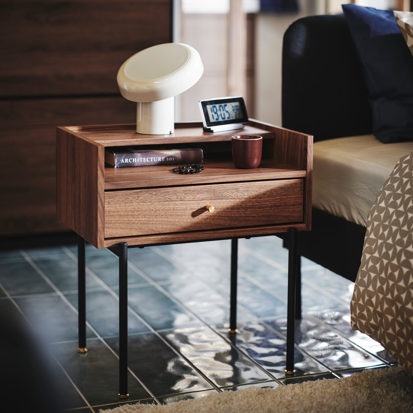 Close-up of a brown walnut-effect RÅDMANSÖ bedside table with a clock, cup book and BLOMPRAKT Bluetooth speaker lamp on top.