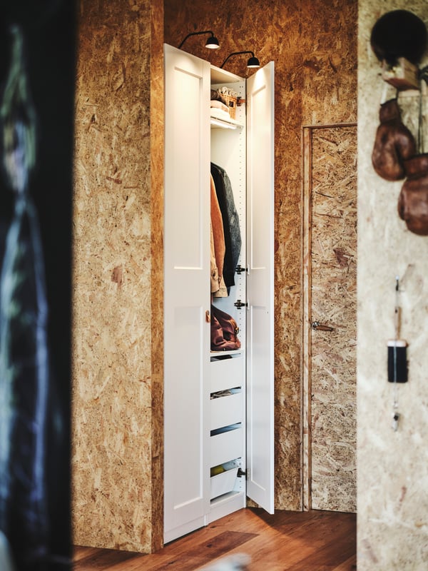 A PAX wardrobe sits in an alcove in a hallway with wood floors. One door is open showing drawers and clothes hanging inside.