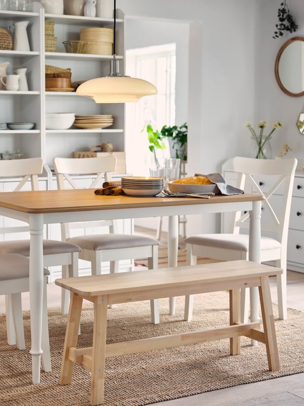 A table in oak/white, four white chairs and a bench in birch stand on a jute rug. Two high cabinets are in the background.