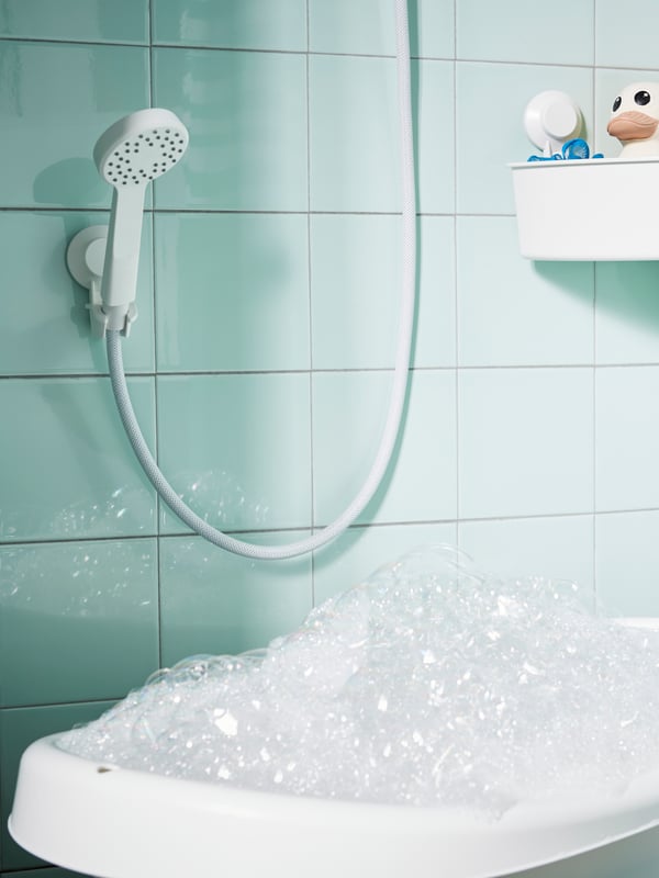 A tiled shower wall with a LILLREVET single spray hand-shower in white and a white TISKEN corner shelf unit with suction cup.