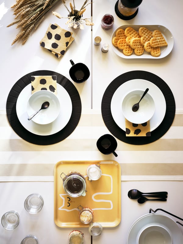 طاولة مع مناديل ورقية HAJMAL باللونين البيج والأسود، وسلطانيات وأطباق ‪IKEA 365‬​​​​​​​ وصينية HAJMAL بنقش قطة.