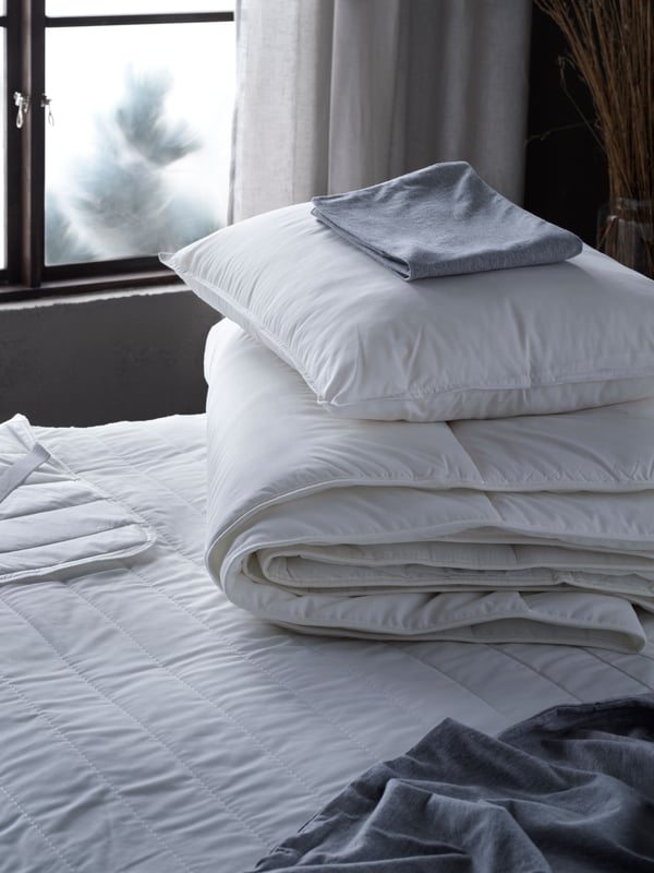 Una colcha blanca LUDDROS, sobre esta se enceuntra una pila de ropa de cama, un plumon blanco SMÅSPORRE abajo de una almohada blanca VILDKORN.