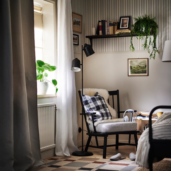 Angle d’une chambre avec fauteuil PERSBOL et lampadaire KLÖVING noir au centre. Fenêtre à gauche et lit à droite.