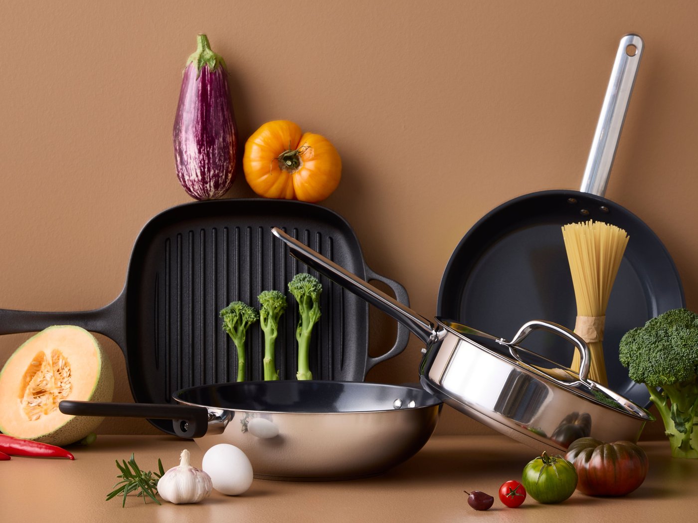 Four pans of different styles and materials are placed on a brown surface with different kinds of vegetables around them.
