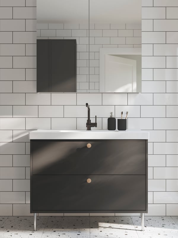 A bathroom with white tiles and a dotted floor has a dark grey HAVBÄCK wash-stand on legs and a mirror cabinet with doors.