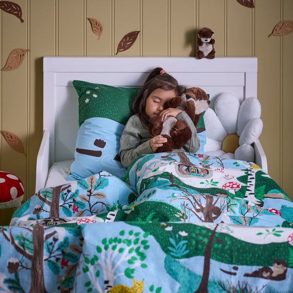 Ein Kind liegt in einem weißen Bett, dessen Bettzeug mit SKOGSDUVA Bettwäsche bezogen ist. Das Kind hält ein SKOGSDUVA Stoffspielzeug in Otterform fest.