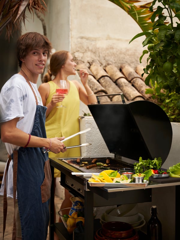 A person wearing a blue/brown GRILLTIDER apron standing in front of an open GRILLSKÄR charcoal barbecue with side table.