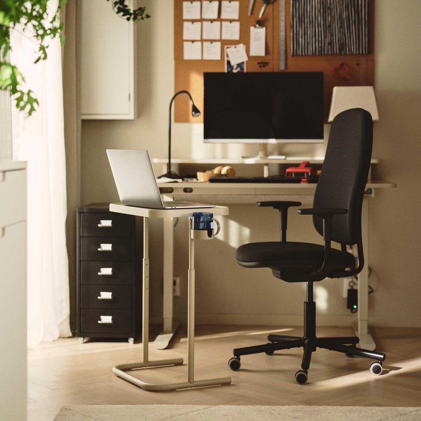 Laptop stand next to a dark grey SMÖRKULL office chair with armrests beside a TRAVSPORT sit/stand desk.