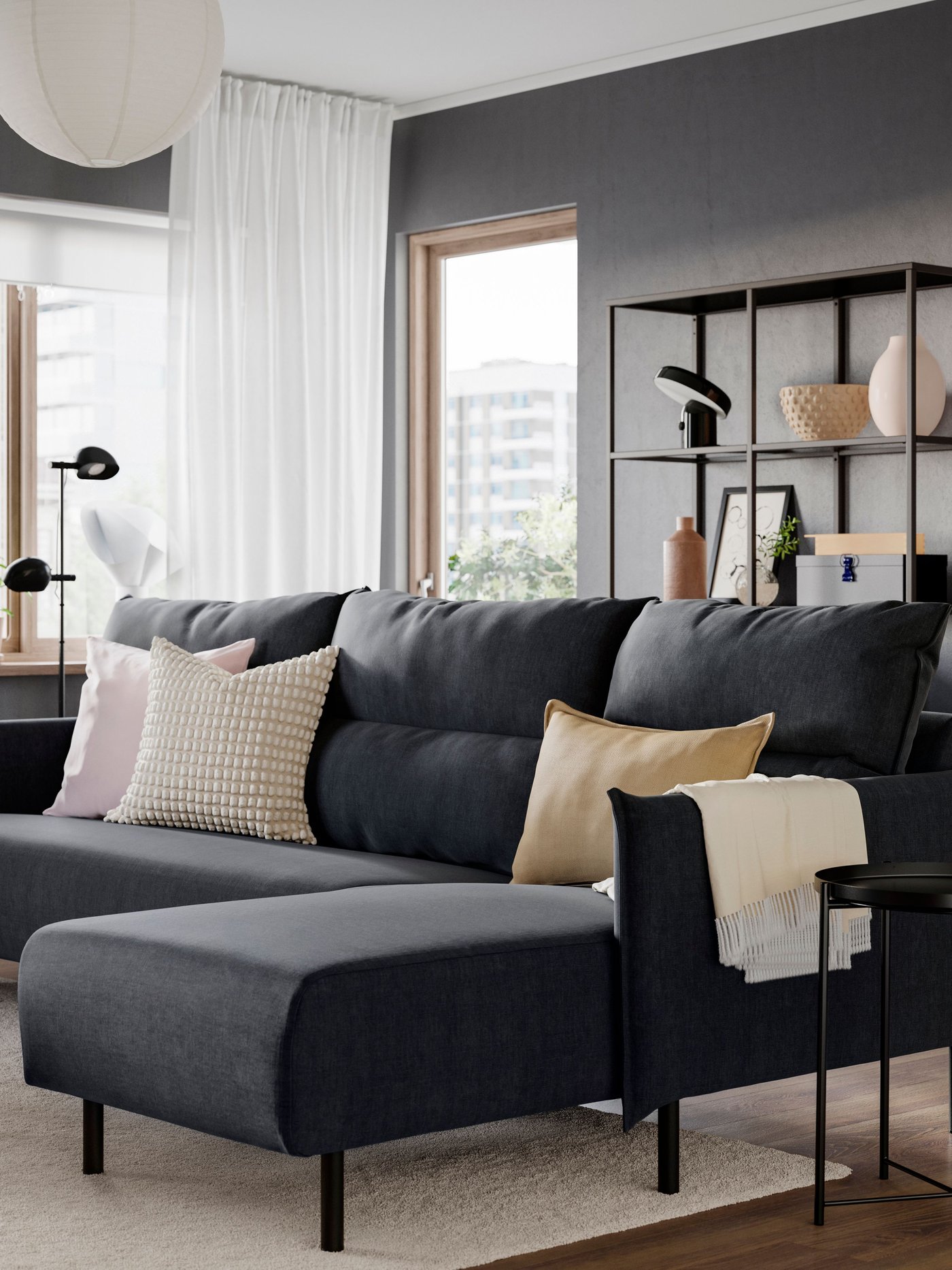 Modernt vardagsrum med mörkgrå väggar och en grå MANNARP soffa med schäslong med ljusa kuddar, placerad vid ett soffbord och stora fönster med vita gardiner.