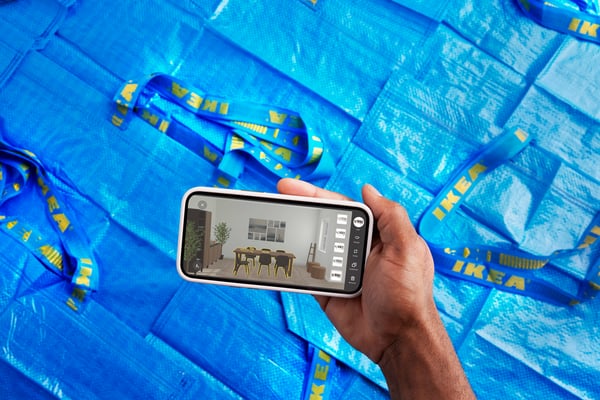 Prikaz izbliza ruku osobe koja drži nagnuti pametni telefon na kojem je prikazana KREATIV funkcija IKEA aplikacije, u kojoj vizualizira nameštenu dnevnu sobu.