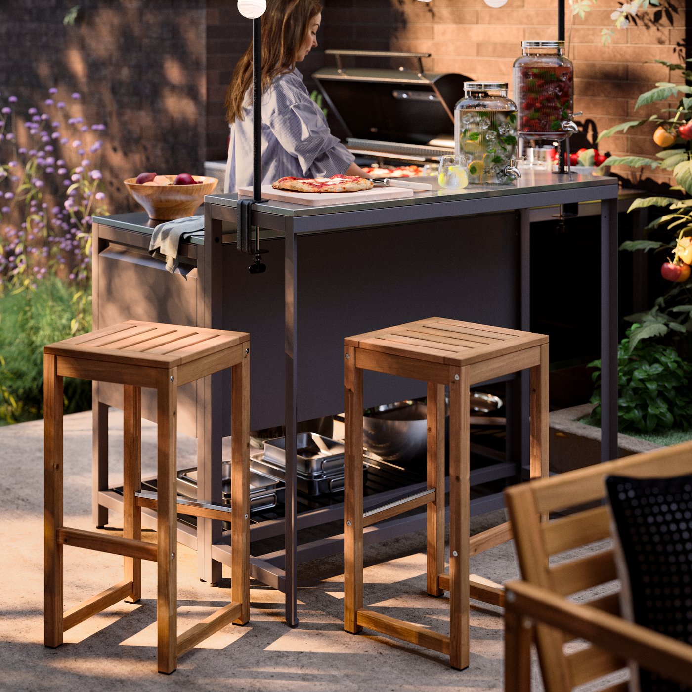 Dos taburetes de bar NÄMMARÖ junto a una cocina exterior en una terraza, donde una persona está cocinando.