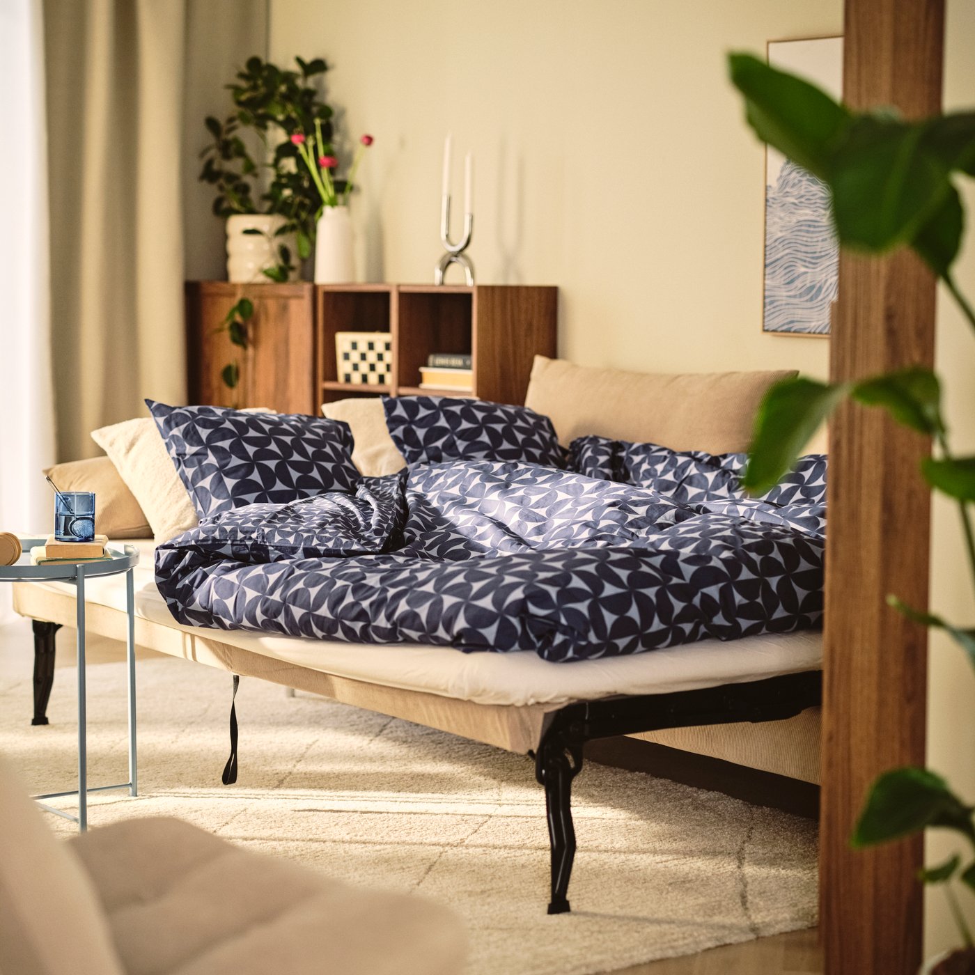 Ein Wohnzimmer mit einem SÖDERHAMN 3er-Bettsofa mit Bezug „Kelinge“ in Beige, umgeben von Kissen und Pflanzen.
