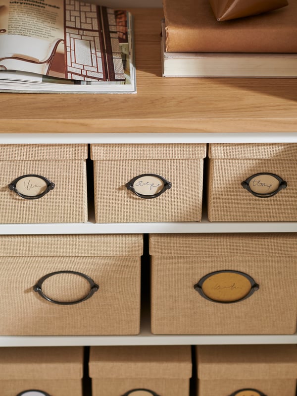 Des étagères BESTÅ avec des livres et un magazine posés dessus, et plusieurs boîtes KVARNVIK beiges avec couvercles, de différentes tailles, rangées à l’intérieur.