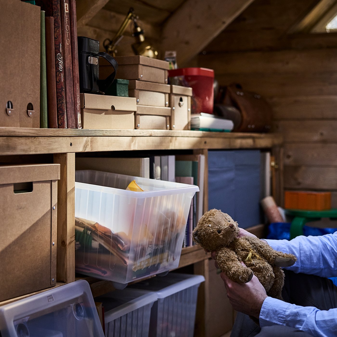 A well-loved teddy bear is being placed in a VESSLA storage crate with castors in an attic storage space.