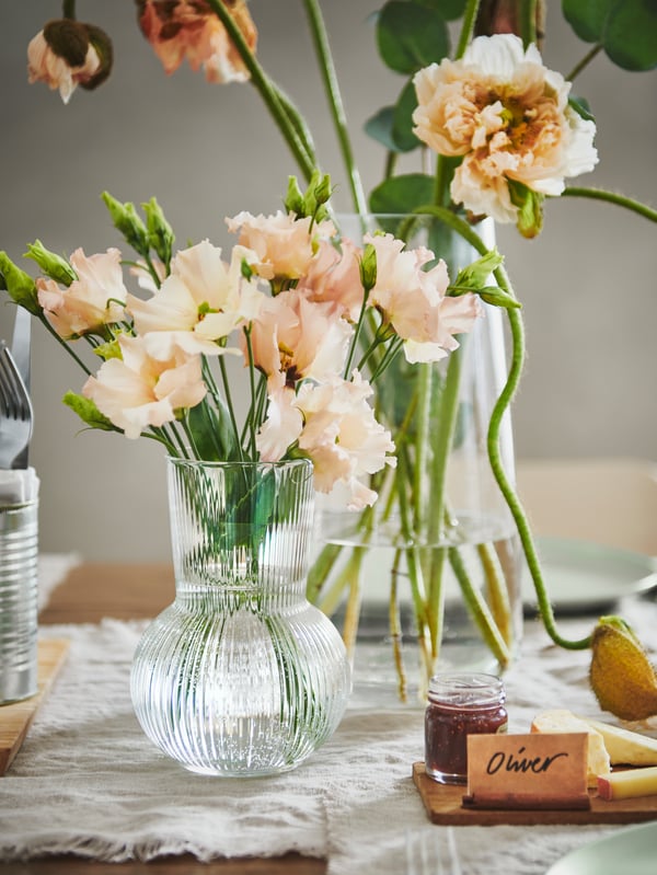 A section of a festively set table with colourful flowers placed in side-by-side PÅDRAG and BERÄKNA clear-glass vases.
