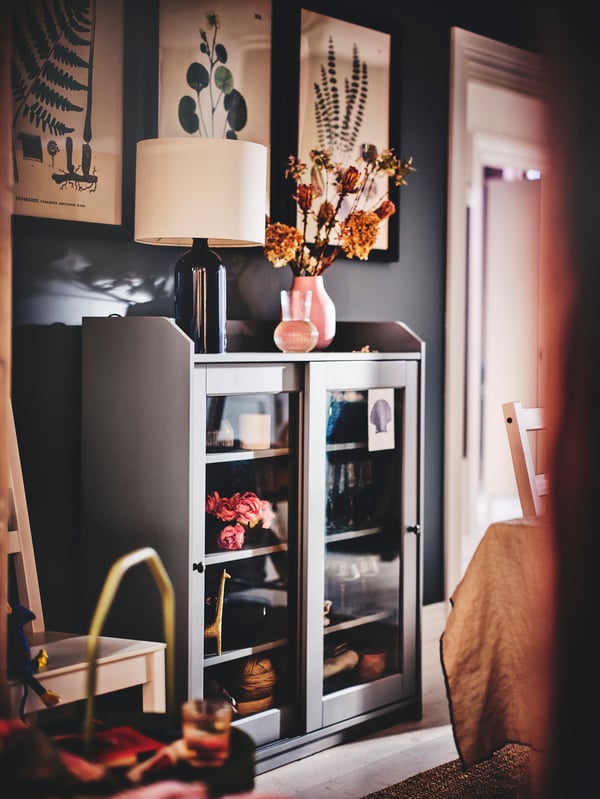 In a dining room is a grey HAUGA glass door cabinet. On the top is a table lamp and a vase with flowers. Inside deco objects.