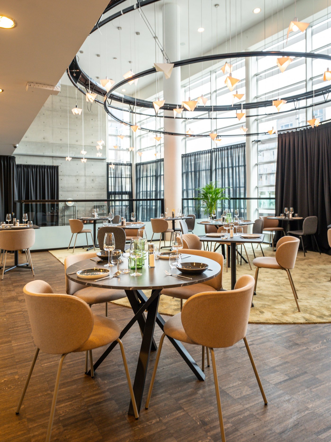 Ronde tafels met beige stoelen in licht restaurant met houten vloer. Grote ramen, zwarte gordijnen en hanglampen in vorm van vogels zorgen voor sfeer.