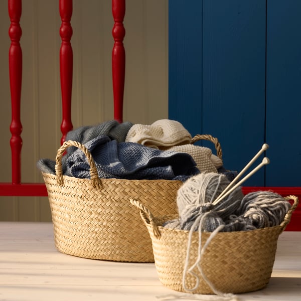 A close-up of a natural woven SANDJORD basket filled with various yarns, set against a blue wall and a red railing.