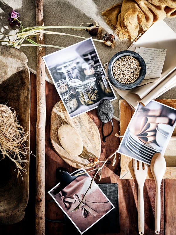 A rustic wooden table top with dried flowers, photographs, straw, fabric, and a wooden spoon, fork and chopping board on top.