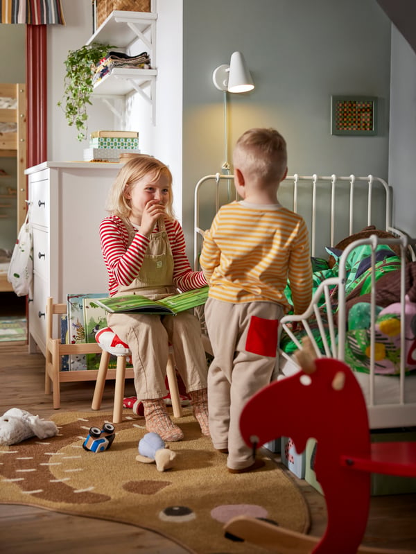 Zwei Kinder befinden sich neben einem ausziehbaren MINNEN Bettgestell. Eines von ihnen sitzt auf einem Hocker und liest ein Buch. Im Vordergrund steht ein EKORRE Schaukelelch.