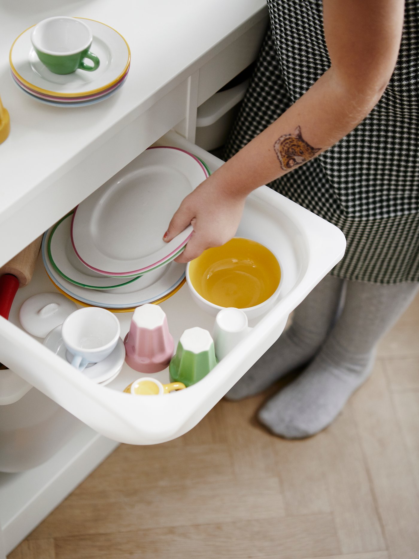 Tiroir ouvert avec assiettes et bols à hauteur d’enfant, main prenant une assiette, tasses et assiettes colorées soigneusement empilées dans une armoire blanche posée sur un plancher en bois.