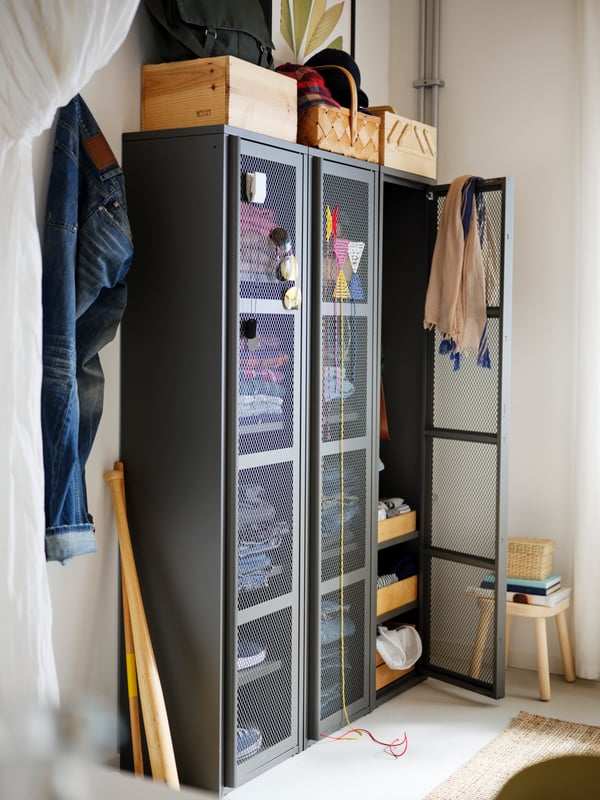 A corner of a room with three high, grey IVAR cabinets with mesh doors filled with clothes, and various boxes on top.