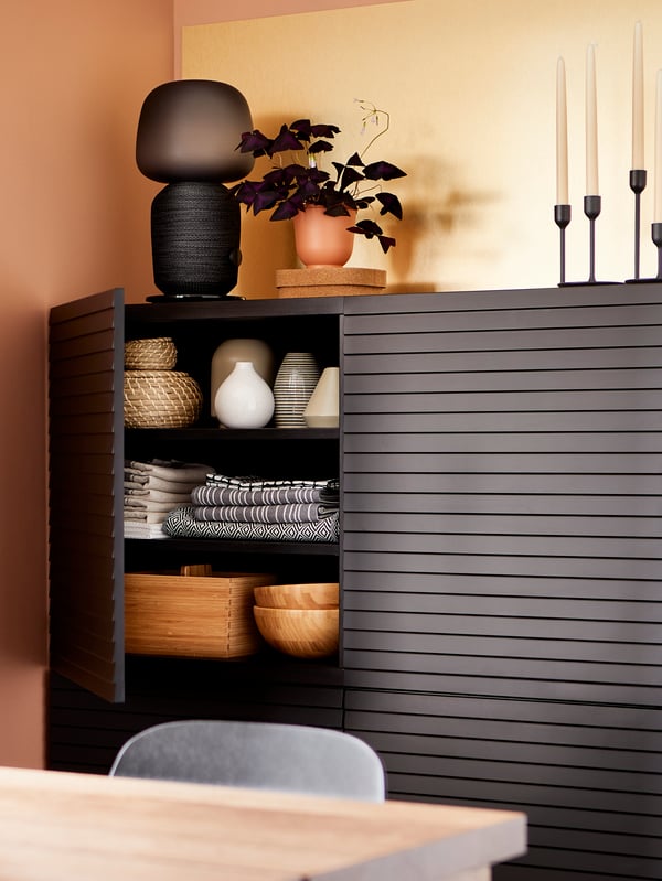 Dining room with storage behind anthracite doors, a brass-coloured wall panel, a black lamp, candles and a purple plant.