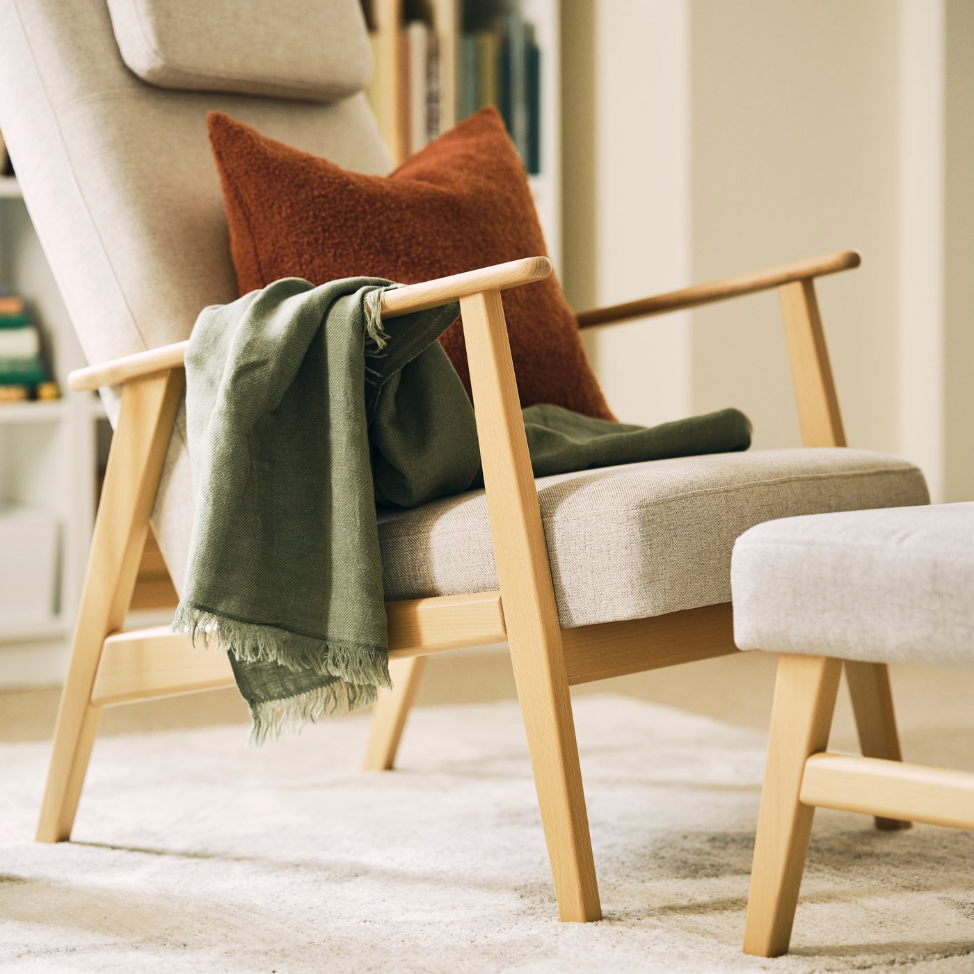A dimly sunlit space with a light-beige EKENÄSET high-back armchair and a footstool, both placed on a light rug.