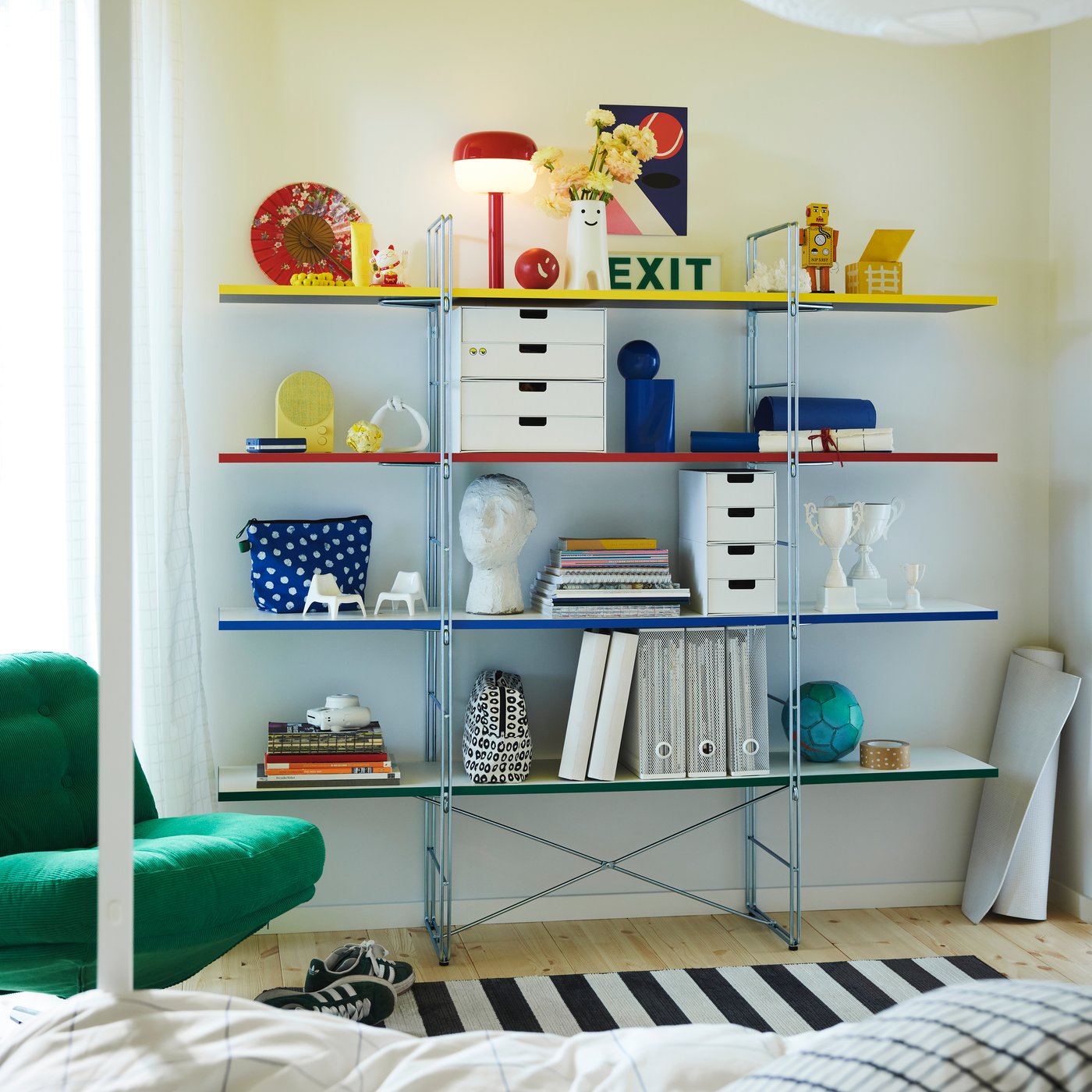 The colourful BYAKORRE open shelving unit holding coloured products such as a lamp, speaker, folders, books and mini chests.