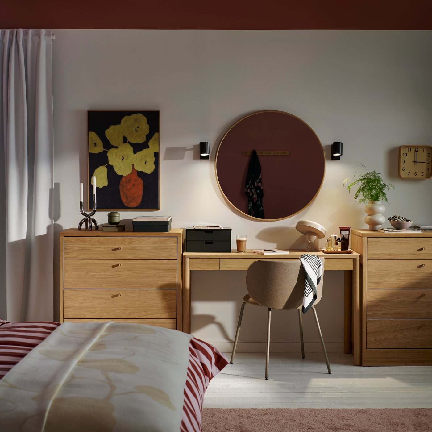 A bedroom with a desk between two oak veneer TONSTAD chests of 4 drawers with items on top and a round mirror on the wall.
