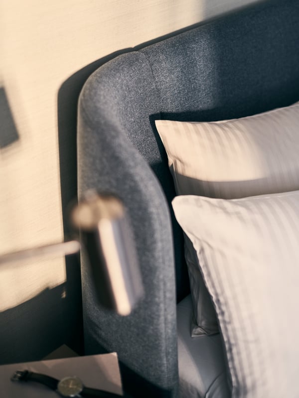 The corner of a blue upholstered TUFJORD bed with two pillows covered in white bed linen, and a silver reading lamp.