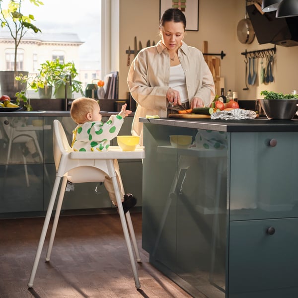 A baby sits in an ANTILOP highchair beside a kitchen worktop wearing a white/green KLADDIG long-sleeved bib and reaching out.