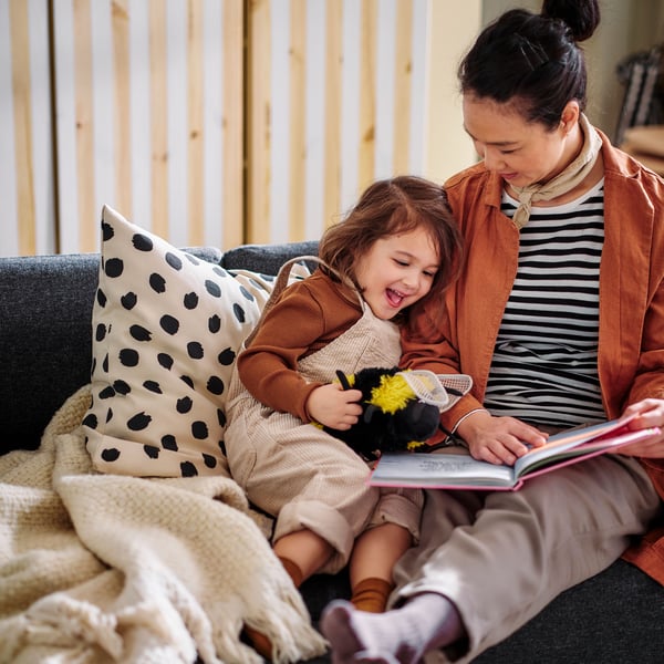 A person reads a story to a small child who is leaning against a cushion with an ODDNY cushion cover on a sofa.
