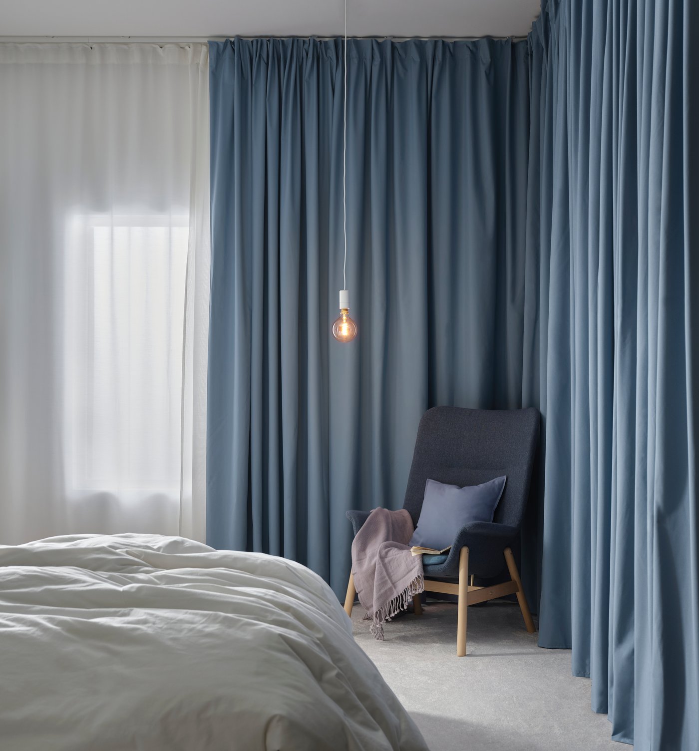 In einem Raum steht ein blauer Stuhl in der Ecke. Davor steht ein weißes Bett mit Bettwäsche und auf der linken Seite sieht man ein Fenster.