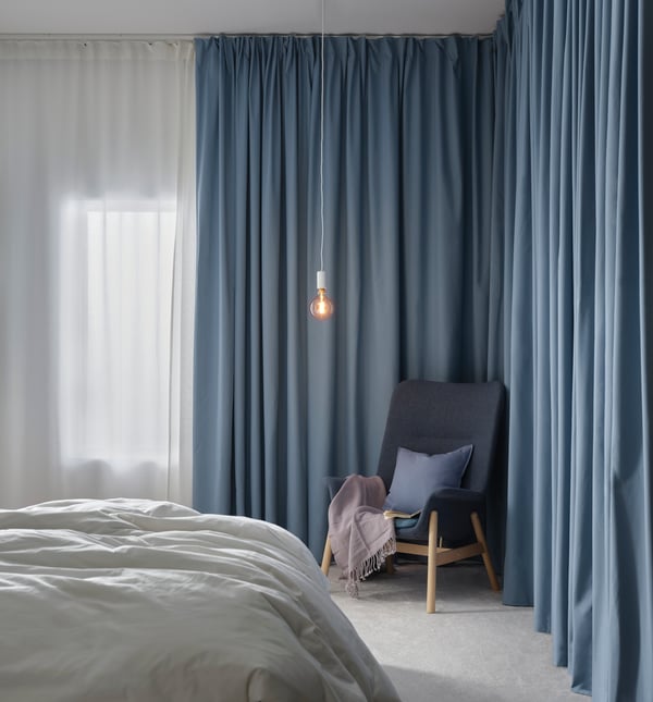 Bedroom with a bedframe in the middle with windows framed with HILJA sheer curtains and BENGTA block-out curtains in blue. A SUNNERBY bulb with cord hangs in the corner above the VEDBO grey armchair with GURLI cushion case on top. 