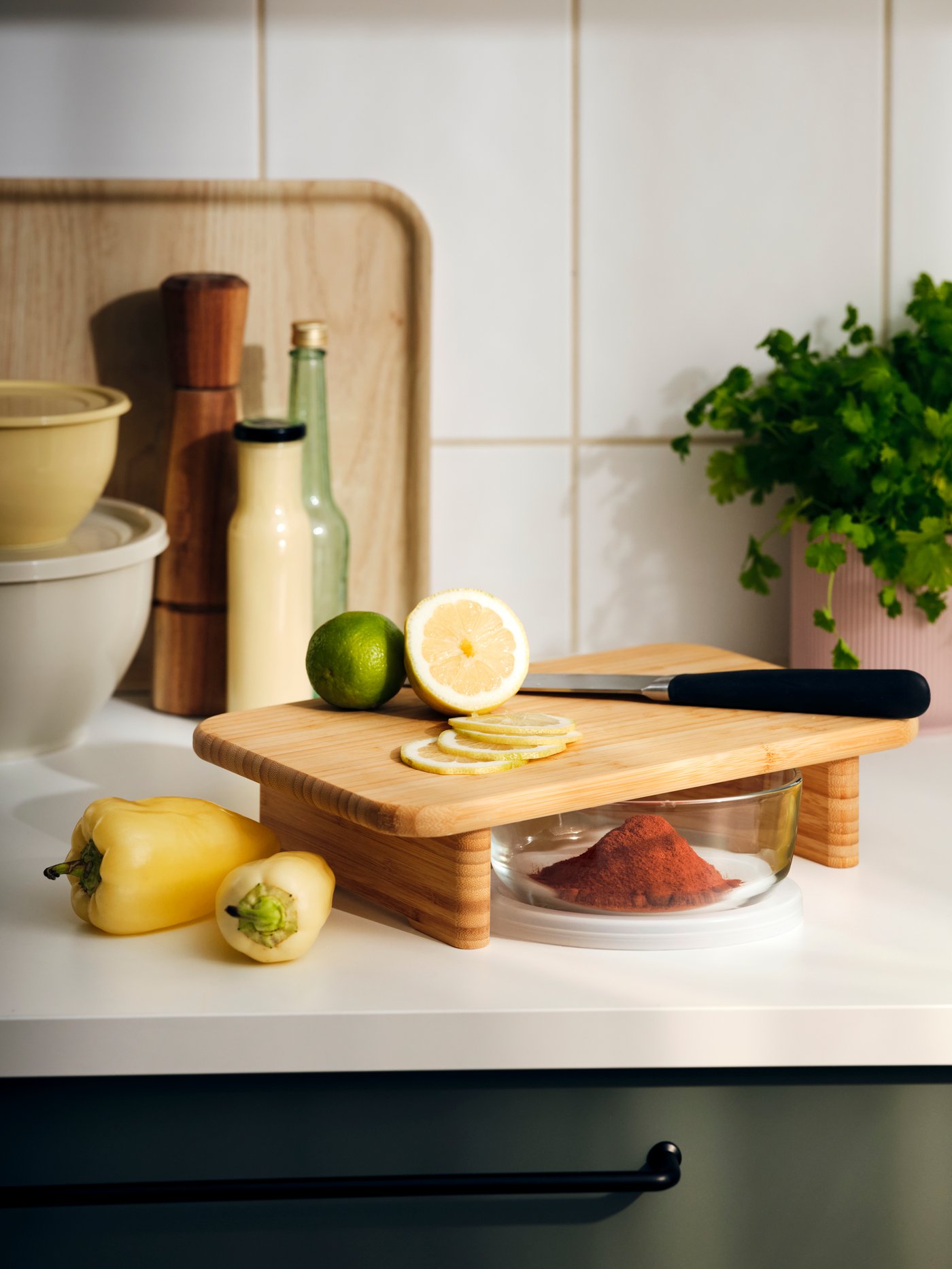 STOLTHET chopping board is on a kitchen counter with a lime and sliced lemon on top; a glass dish is in the space underneath.
