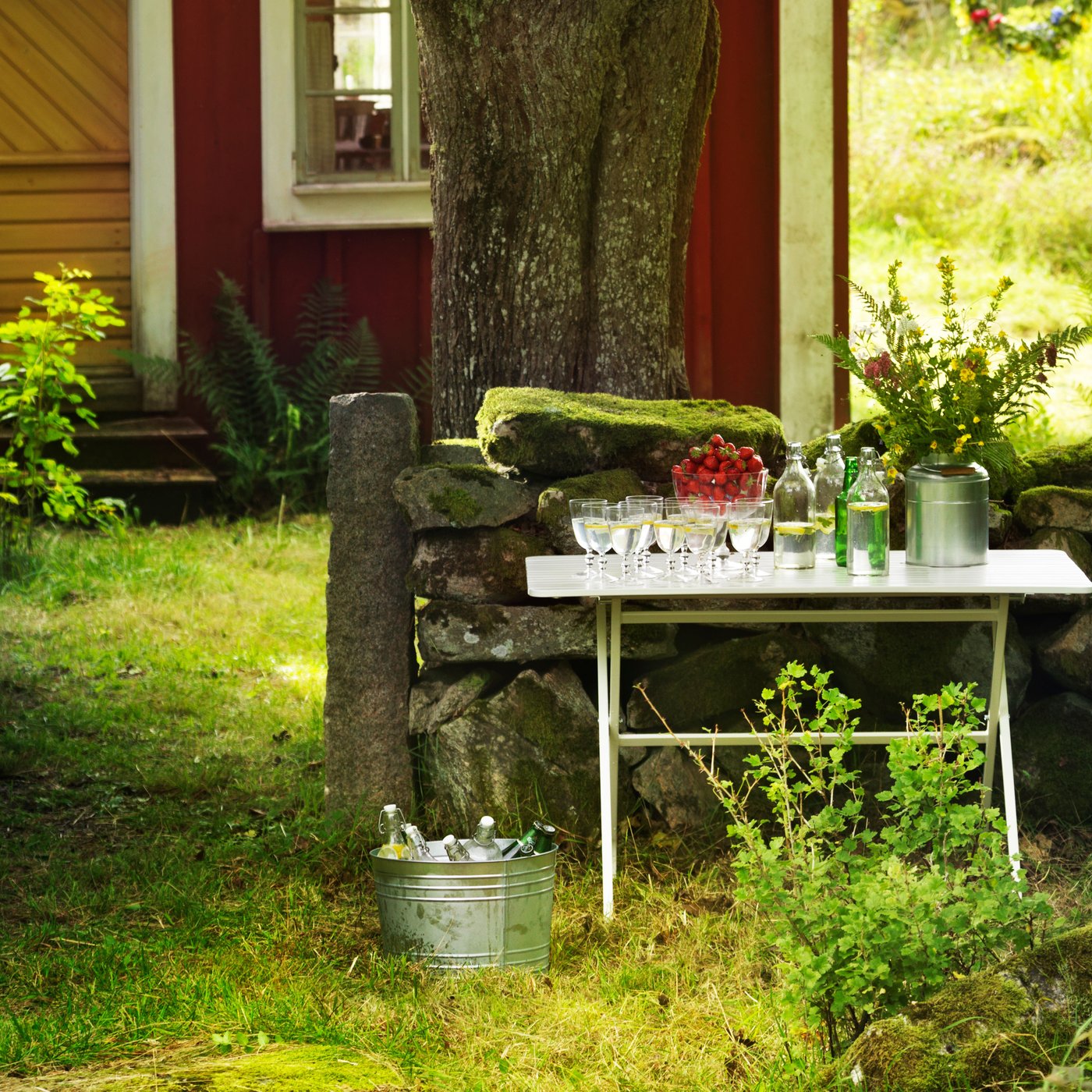Kleiner Gartentisch mit Getränken, Gläsern und Blumen vor einem alten Steingarten und einem roten Holzhaus im Grünen.