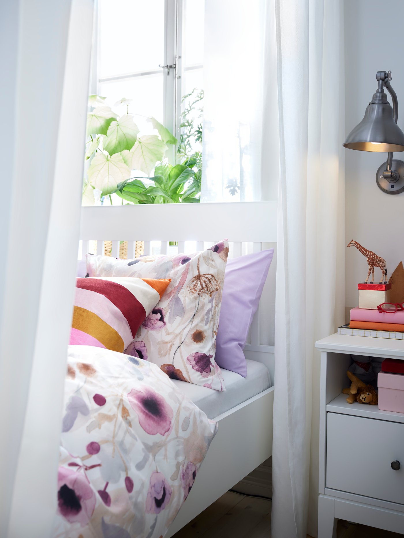 Ein weißes IDANÄS Bett mit LÖNNHÖSTMAL Bettwäsche steht hier neben einem IDANÄS Ablagetisch unter einem Fenster.