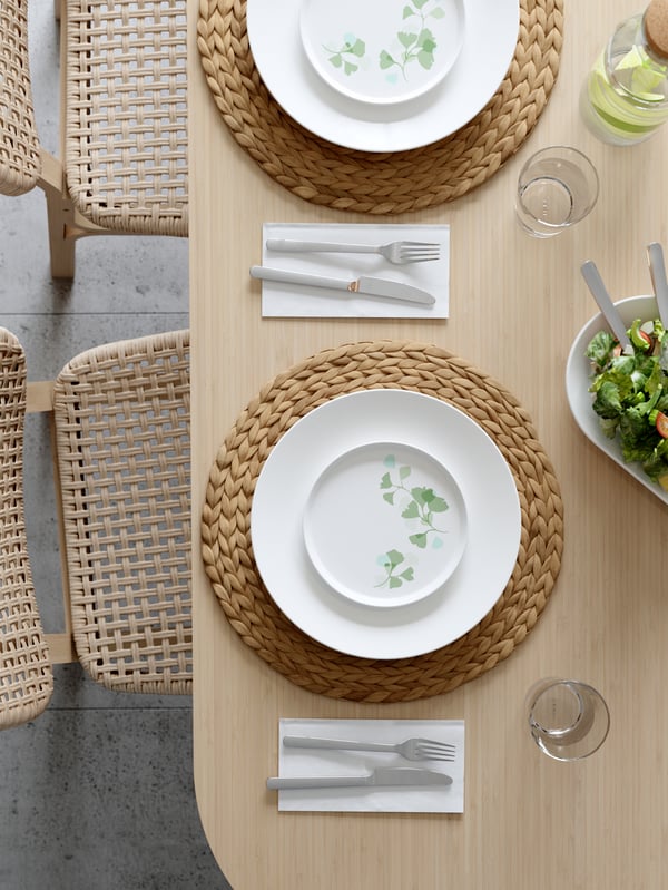 Tisch für zwei Personen gedeckt mit gewebten Tischsets, Tellern mit Blattmotiven, Gabeln und Messern auf Servietten, Gläsern und einer Salatschüssel auf einer hellen Holzfläche.