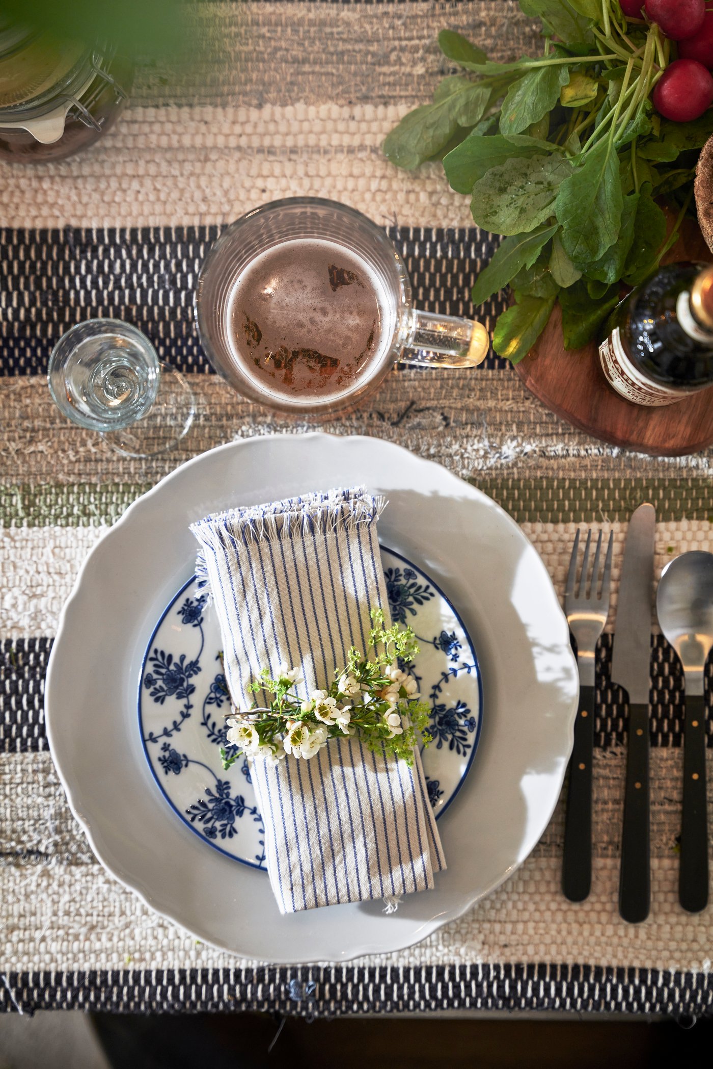En elegant bordsdukning sedd uppifrån, med en blåmönstrad tallrik som toppas av en randig servett och färska blommor. En mugg med öl och bestick med mörka handtag är placerade på en vävd, randig bordstablett.