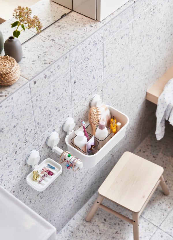 Vista superior de canastos de baño TISKEN blancos, adheridos con ventosas a un muro con baldosas con diseño de terrazzo en tonos grises. Bajo los canastos hay un piso de madera VILTO. 