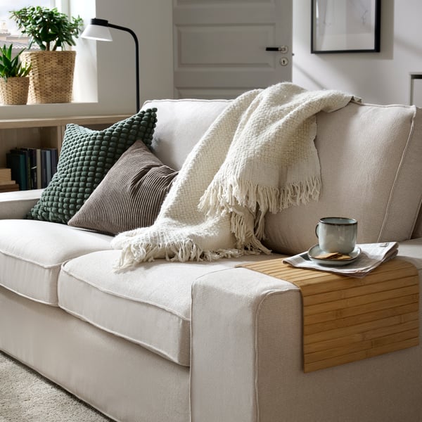 A light living room with a two-seat KIVIK sofa with cushions, a throw, and a coffee cup on a RÖDEBY armrest tray on one end.