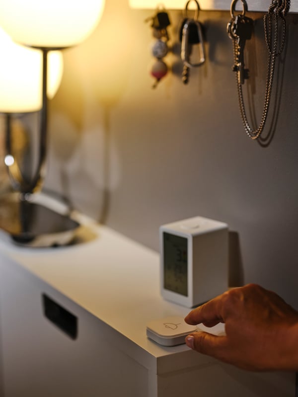 A hand touching a TRÅDFRI shortcut button that’s on top of a white STÄLL shoe cabinet alongside a SIMRISHAMN table lamp.