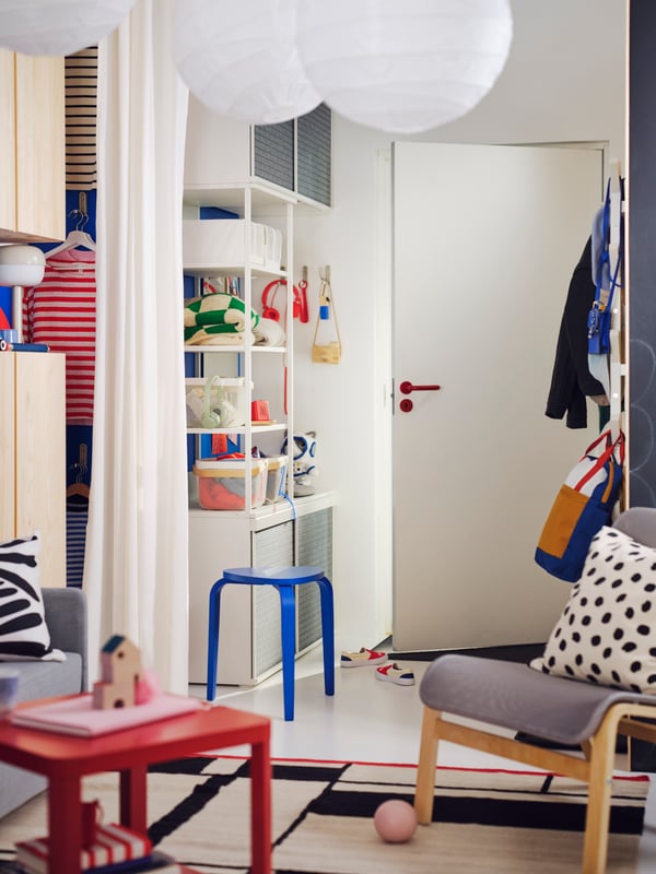 A PLATSA open wardrobe with sliding doors by a front door holds clothes, toys and headphones. A blue KYRRE stool is nearby.