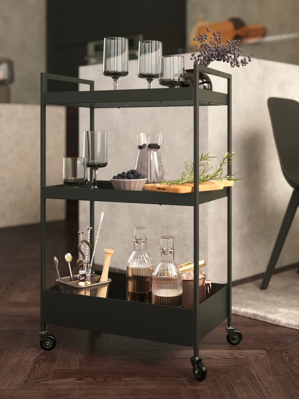 A NISSAFORS cart in black holding OMBONAD wine glasses and goblets in grey, carafes, a chopping board and other items.