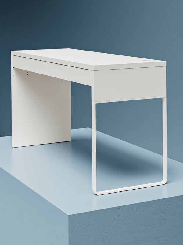 A space with semi-lit, unfurnished walls and a low podium with a single, white MICKE desk placed on it.