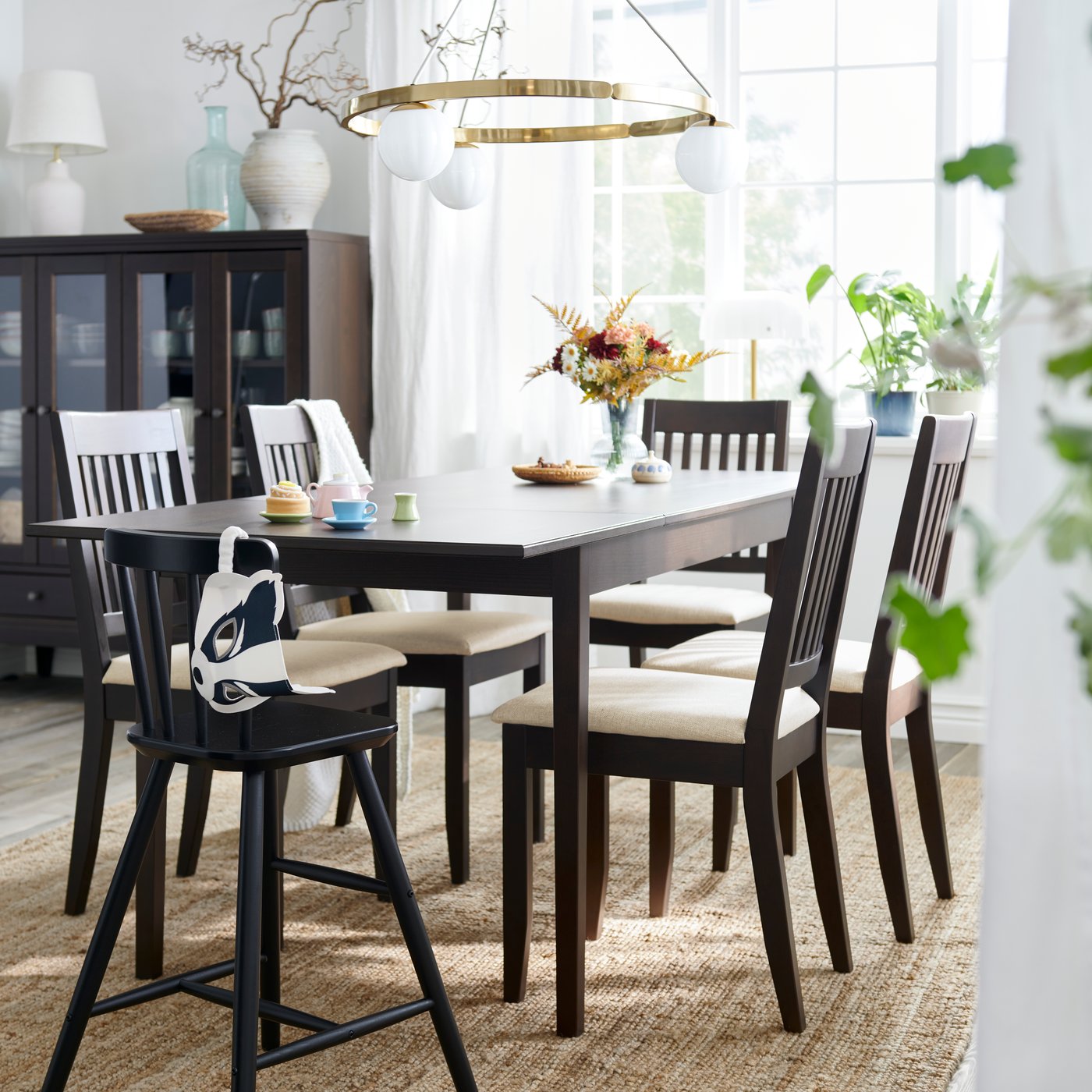 Table extensible NÄSINGE en brun foncé et chaises dans une salle à manger lumineuse dotée de grandes fenêtres et agrémentée de plantes vertes.