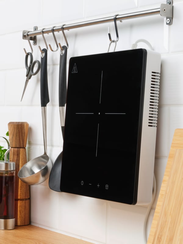 A TILLREDA portable induction hob 1 zone in white, hanging on a rail on the wall, along with scissors and kitchen utensils.