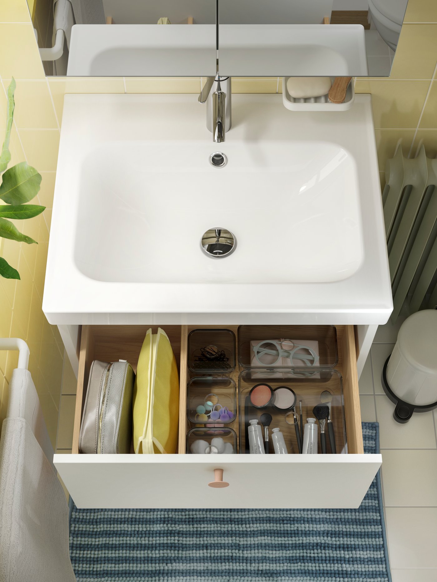 Un meuble de salle de bain avec tiroir ouvert présente un rangement soigné de produits de toilette, maquillage et soins, avec un miroir au-dessus et un tapis rayé bleu au sol.