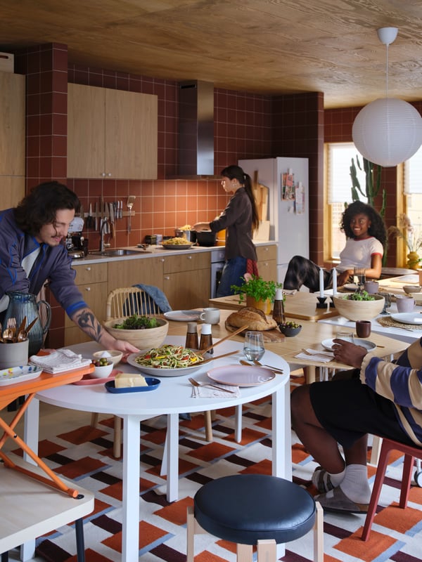 Four people are preparing for a meal in a lively kitchen. One person leans over a white VIHALS table, placing food on it.